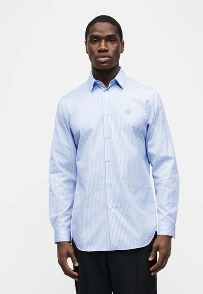 Μαύρος άνδρας που φοράει γαλάζιο και λευκό ριγέ πουκάμισο με κουμπιά και μακριά μανίκια και μαύρο παντελόνι, στέκεται μπροστά σε απλό φόντο.