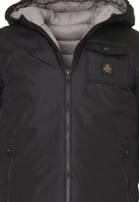 Giubbotto puffo nero con cappuccio staccabile, zip sul davanti e tasca sul petto con un patch logo. Texture esterna liscia, foderato isolante.