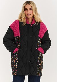 Čierna prešívaná bunda s ružovými textúrovanými ramenami, bočnými časťami s kvetinovým vzorom a zapínaním na cvoky. Má manžety na rukávoch a bočné vrecká.