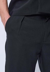 Un pantalon noir ajusté présente un tissu lisse, une coupe slim, des poches latérales et un devant légèrement plissé. La ceinture est également conçue de manière discrète.