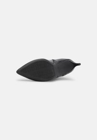 Botín negro de punta afilada con suela de goma texturizada y tirador en la parte trasera, visto desde la parte inferior sobre un fondo blanco.