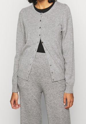 Femme portant un cardigan gris clair boutonné et un pantalon en maille assorti par-dessus un haut noir, debout devant un fond blanc uni.
