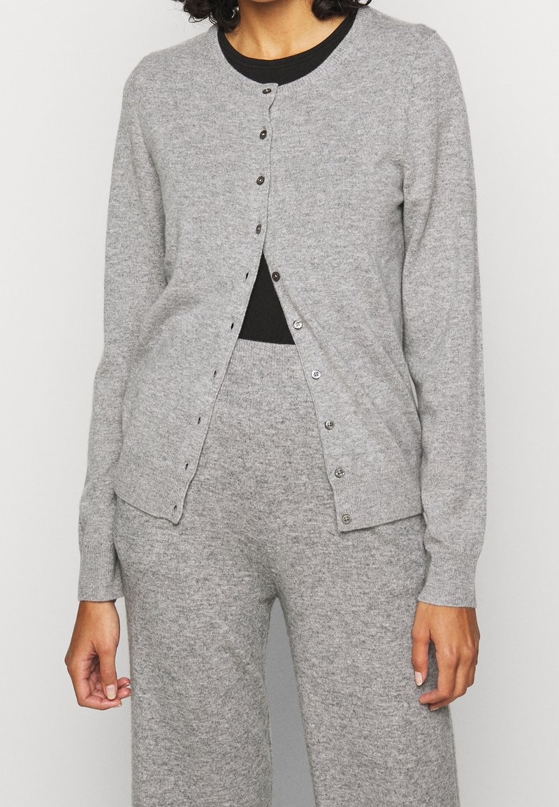 Femme portant un cardigan gris clair boutonné et un pantalon en maille assorti par-dessus un haut noir, debout devant un fond blanc uni.