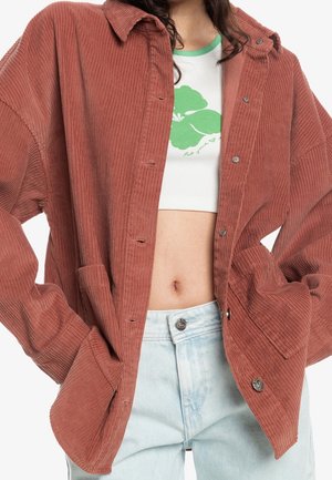 Osoba ubrana w otwartą, rdzawe sztruksową kurtkę na białym krótkim topie z zielonym kwiatowym wzorem i jasnoniebieskie, z wysokim stanem dżinsy, ręce w kieszeniach.