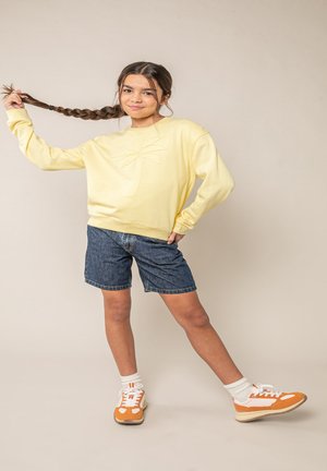 Jonge vrouw die een gele trui met palmboomprint draagt, spijkershorts en oranje sneakers, terwijl ze haar gevlochten haar vasthoudt en poseert.