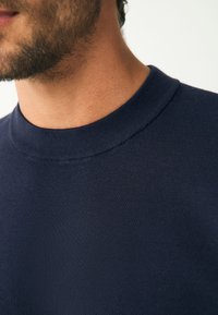 Tumši zils džemperis ar apaļu kakla izgriezumu, izgatavots no gludas, adītas auduma, ar cieši pieguļošu ribveida apkakli un izsmalcinātu dizainu.