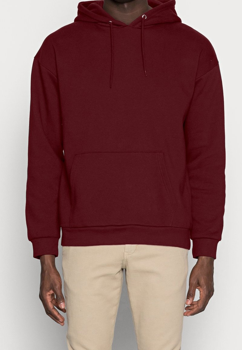 Personne portant un sweat à capuche bordeaux uni avec poche avant et pantalon beige, debout devant un fond clair.