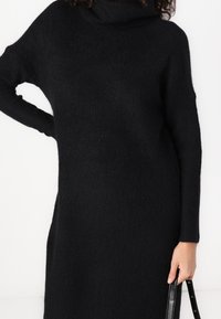 Robe-pull noire en tricot avec un col cheminée, des manches longues, une texture côtelée et des fentes sur les côtés. Accompagnée d'un sac à main noir.