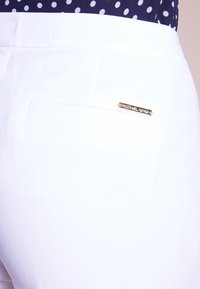 Vita byxor med en slät yta, som har en bakficka och en gyllene "Michael Kors" logotypdetalj. Prickig topp delvis synlig ovanför.