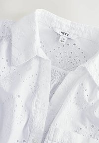 Blouse en coton blanc avec un motif de broderie fine, un col classique et une fermeture à boutons. L'étiquette à l'intérieur indique la marque et la taille.