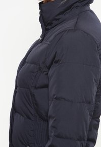 Marineblå dunjakke med høj krave, forreste lynlås, blød tekstur og vandret syninger for isolering.