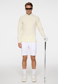 Homme portant un haut à manches longues crème, un short blanc, des gants blancs et des lunettes de soleil, tenant un club de golf, debout devant un fond uni.