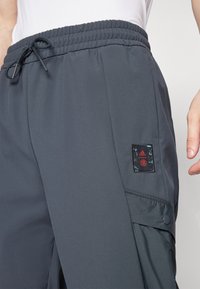 Pantaloni sportivi grigio scuro con vita elastica e cordino, dotati di una tasca laterale con chiusura a pressione e una toppa con il logo Adidas.