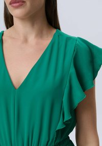 Top de tejido ligero y verde que presenta un escote en V profundo y mangas acampanadas. La textura es suave, con un detalle de cintura ajustada.