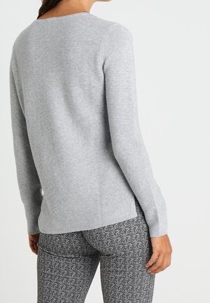 Femme portant un pull côtelé gris clair à manches longues et un pantalon gris motif chevrons, vue de dos et de la taille vers le haut.