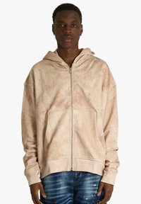 Sweat à capuche beige zippé avec une finition texturée, capuche ajustable et poches avant ; présente une coupe décontractée et des poignets côtelés.
