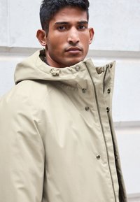 Beige vandtæt jakke med høj krave, justerbar hætte og trykknapper. Glat tekstur, strømlinet design og sølv hardware-detaljer.
