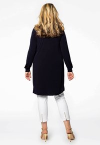 Langermige marine tuniek met een losse pasvorm, gecombineerd met witte cropped pants met zijkleppen, en hoge sandalen.