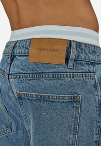 Lähikuva vyötärön takapuolelta, jossa näkyy vaaleansiniset farkut, joissa on ruskea nahkainen "PEGADOR"-merkintä ja valkoinen vyötärönauha alla.