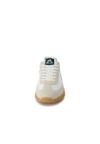 White sneaker with beige suede accents, textured gum rubber sole, and classic lace-up design. Logo on tongue in green and white.