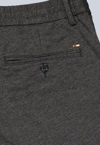 Nahaufnahme von dunkelgrauen, strukturierten Stoffhosen, die eine Gesäßtasche mit einem schwarzen Knopf und einem kleinen rechteckigen Metallschild zeigen.