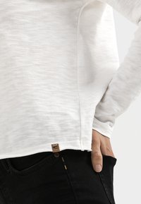 Witte blouse met lange mouwen en een gestructureerde afwerking, met een zijsplit aan de onderkant en een klein bruin merklabel aan de linksonder. Zwarte jeans zichtbaar.