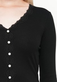 Haut noir à manches longues à rayures, avec un col en dentelle en forme de scallop et un devant boutonné avec des boutons blancs.