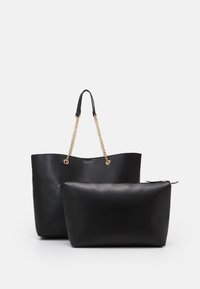 Borsa tote in pelle nera con manici in catena dorata e pochette zippata nera coordinata davanti su sfondo bianco.