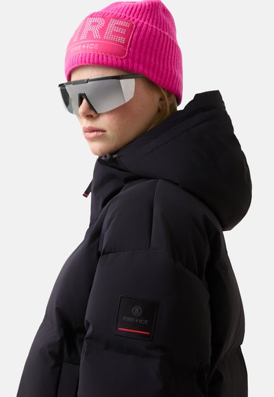 Zwarte pufferjas met capuchon, voorzien van een logopatch op de mouw. Felle roze geribbelde gebreide muts en reflecterende zonnebril maken de look af.