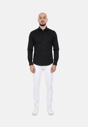 Uomo in piedi, indossa una camicia nera aderente con bottoni, pantaloni bianchi slim e sneakers bianche su uno sfondo chiaro e uniforme.