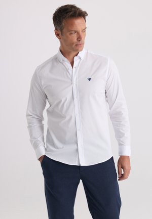 Man wearing a white buttoned shirt with a small blue bird logo on the chest and dark blue pants, standing with hands in pockets.