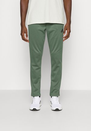 Grüne Herren-Sporthose aus glattem Stoff mit konisch zulaufenden Beinen und seitlichen Nahtdetails, kombiniert mit weißen Sportschuhen.