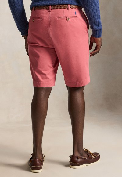 Mann trägt korallrosa Shorts, braunen geflochtenen Gürtel, blauen Pullover und braune Leder-Bootsschuhe und steht vor neutralem Hintergrund.