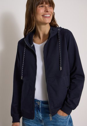 Femme souriante, portant un sweat à capuche zippé bleu marine avec cordons sur une chemise blanche et un jean bleu, la main droite dans la poche.