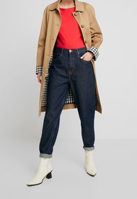 Trench beige con fodera a quadri, t-shirt rossa, jeans scuri a vita alta, e stivaletti bianchi a punta con tacco a blocco.