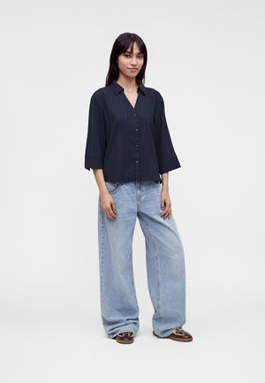 Jeune femme aux cheveux raides et foncés porte une chemise bleu foncé à boutons avec de larges manches trois-quarts et un jean bleu clair ample avec des chaussures marron.