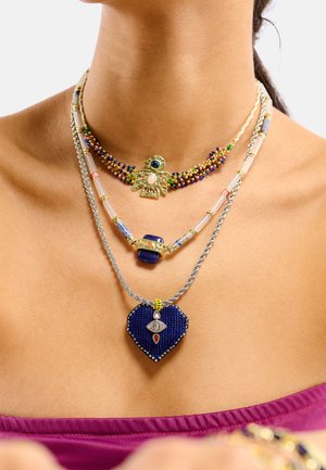 Collier à plusieurs rangs avec perles colorées, pendentif oiseau en or, et charme en forme de cœur bleu avec des détails complexes et des accents en argent.