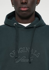 Sweat à capuche vert foncé avec un tissu texturé, arborant un grand logo brodé "ORIGINALS". Il possède une capuche avec cordon et une coupe décontractée.