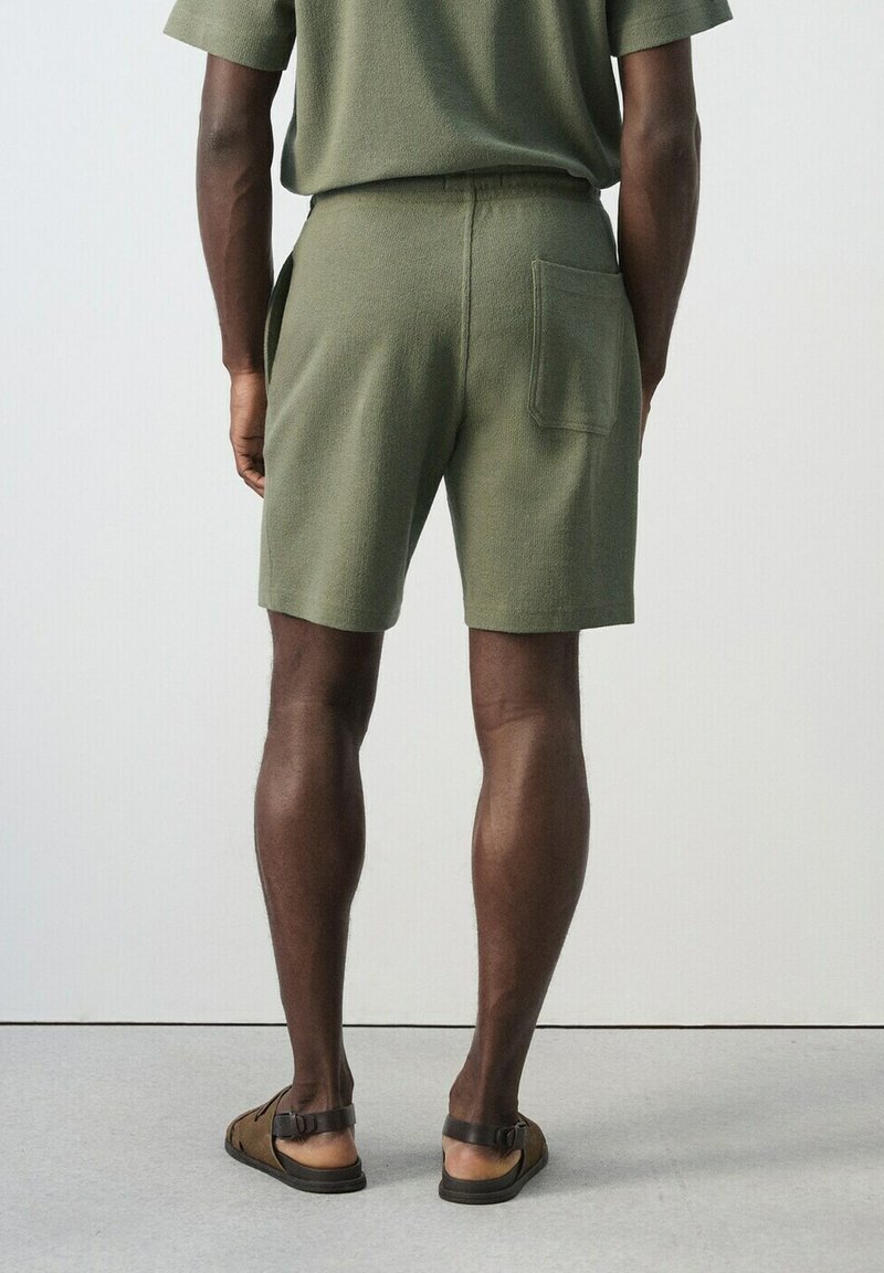 Homme portant un short en maille vert olive et une chemise assortie, debout de dos, avec des sandales marron sur un sol gris.