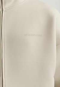 Sudadera con cremallera en un beige claro, hecha de un tejido suave con acabado mate. Presenta un logo en relieve en el pecho y una cremallera texturizada.