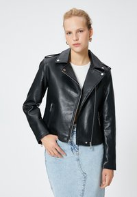 Svart läderbikerjacka med silverdetaljer, sned dragkedja och två dragkedjefickor. Den bärs över en vit topp och en ljusblå denimkjol.