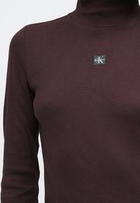 Bordowy prążkowany top z golfem o dopasowanym kroju oraz czarną kwadratową naszywką z białym napisem na górnej części klatki piersiowej.