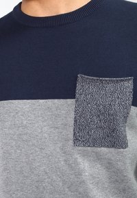 Pull coloré bloc en bleu marine et gris avec une poche en patch texturé grise, un col côtelé et un tissu en tricot doux.