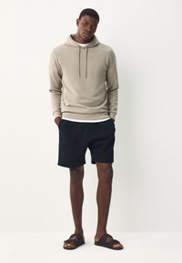 Beige hoodie with drawstrings, paired with black textured shorts and black sandals. Model stands with hands in pockets, neutral background.