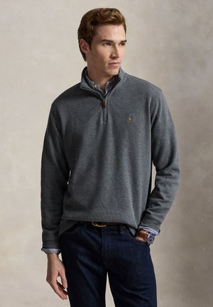 Jeune homme portant un pull gris foncé à fermeture éclair partielle sur une chemise rayée et un jean foncé, debout avec une main dans la poche devant un fond uni.