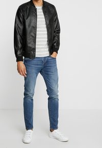 Veste bomber en simili cuir noir, t-shirt rayé gris, jeans bleus et baskets blanches. Design simple avec des poignets et une taille côtelés.