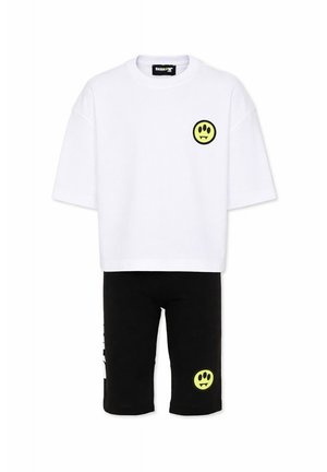 Wit oversized T-shirt en zwarte shorts, elk met een klein geel smileygezicht met hoektanden op de borst en het been.