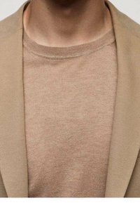 Blazer beige sur un pull léger beige à col rond, présentant une texture lisse et des lignes épurées avec des coutures subtiles visibles à l'épaule.
