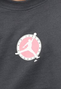 Σκούρο γκρι ύφασμα με λευκή σιλουέτα ενός άλτη μπάσκετ πάνω σε ροζ κύκλο, περιβαλλόμενο από το κείμενο «Air Jordan Flight Club».