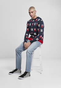 Pulover tricotat cu design de Crăciun în nuanță de bleumarin, având fulgi de zăpadă roșii și albi și figuri de Moș Crăciun. Asortat cu blugi de culoare albastru deschis și adidași cu carouri negre.